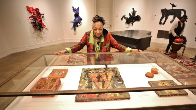 A Black artist, Barbara Bullock, smiles and leans over, touching a glass case containing some of her small works in a large retrospective exhibit at Woodmere Art Museum.