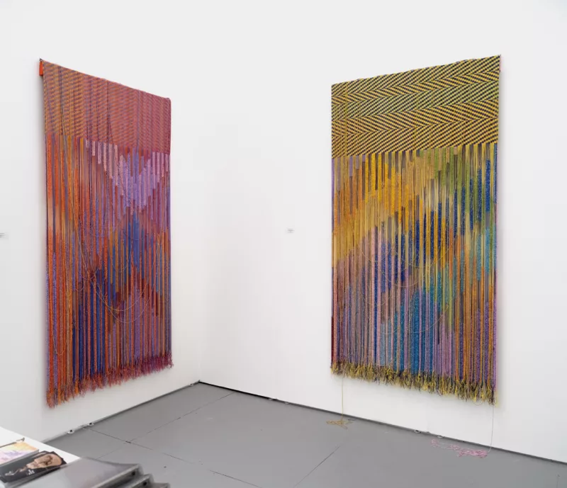 A corner of an art gallery booth at a Miami art fair shows two partially woven pieces whose unwoven threads hang down almost to the floor.