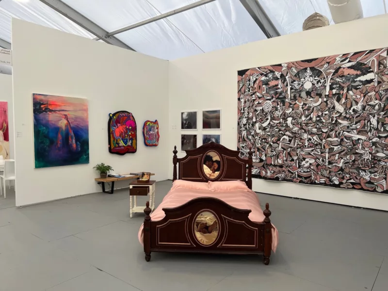 A corner of a gallery booth at a Miami art fair shows colorful paintings on the walls and in the center of the space, a large ornate bed with dark headboard and footboard, pink silk bed linens and what looks like color photos of a black couple embracing,