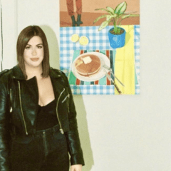 Megan Galardi stands to the left, in front of a wall. To the right hangs a painting of a table top with a plate of pancakes and a potted plant.