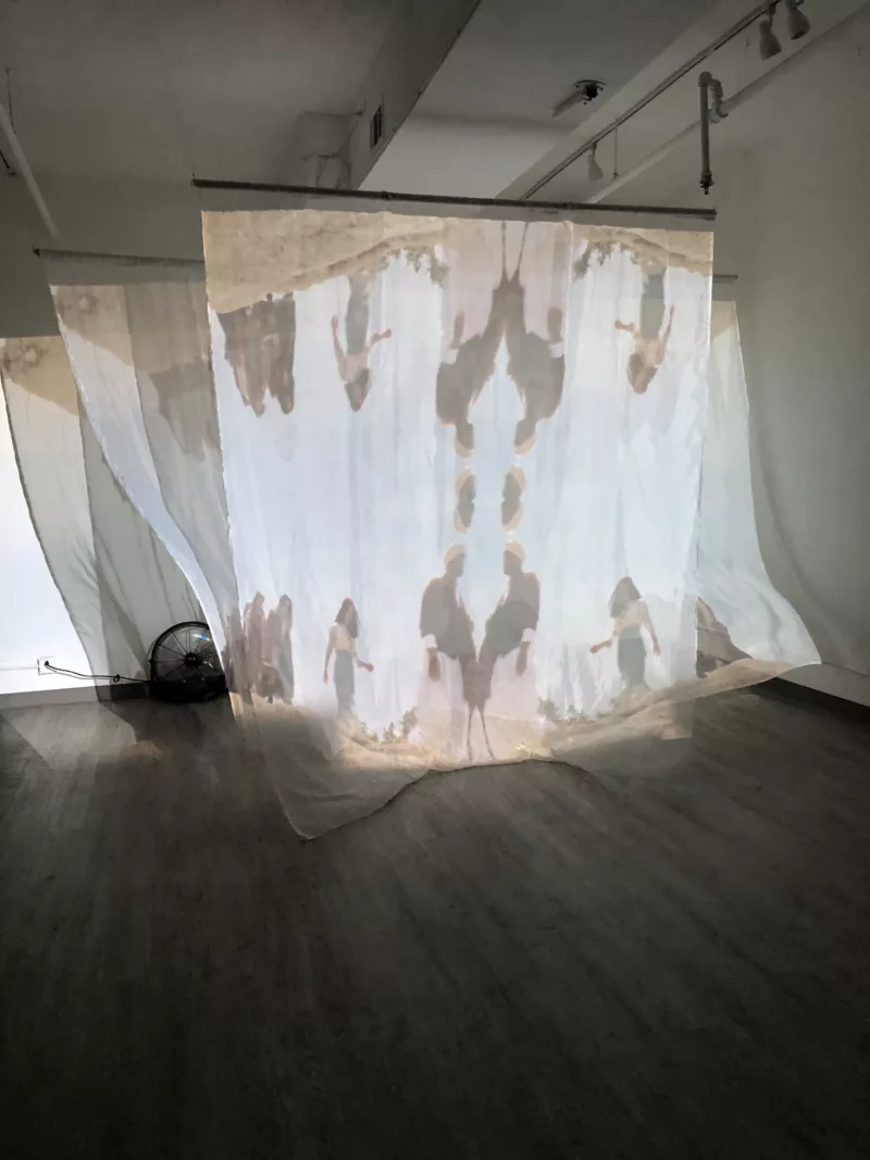 A group of three or more sheer cloth scrims are hung like room dividers in a gallery with a wood floor. A fan plays on them making them move in a ghostly ethereal way.