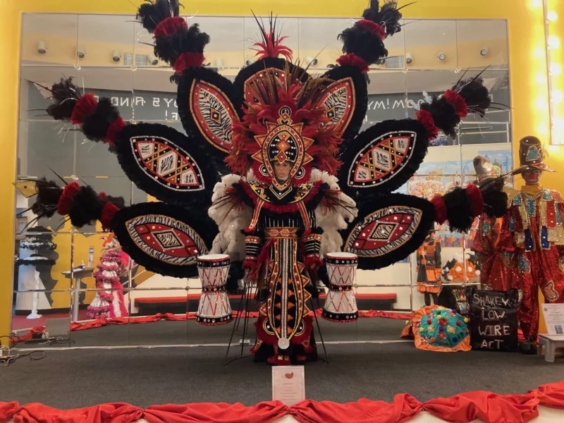 A colorful museum display shows a Mummers costume worn by a manikin, with feathers, sequins, gold touches and to the side of the manikin, two decorated bongo drums.