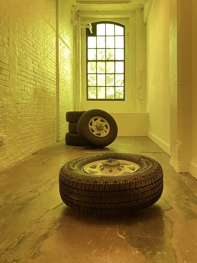 A long narrow gallery is imbued with a garish yellow light, perhaps created by the color of paint on all the walls and around the large window. On the floor are five car tires.