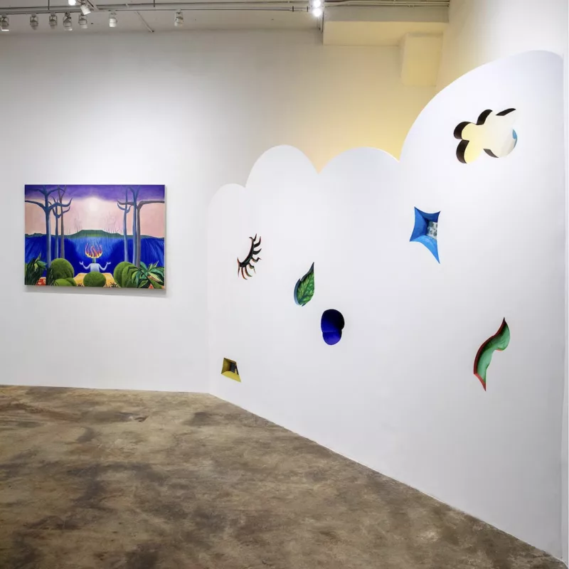 A gallery installation shows a partial wall with a scalloped top edge on the right, with small surreal window-like cut outs into the wall that take the shape and color of a leaf in one instance and in others, something abstract. Behind the scalloped wall is a yellow light emanating.