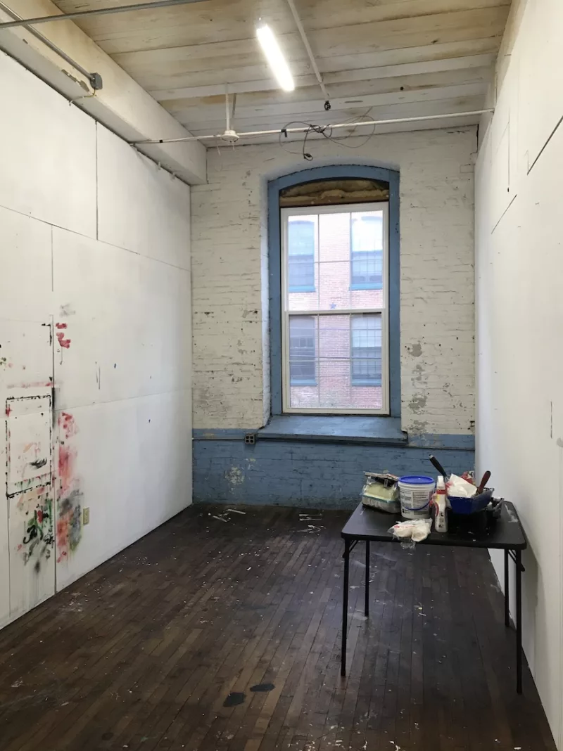 A long narrow room in need of some love and attention. The white paint on the far brick wall is coming off, the left wall has daubs of paint in red and green leftover from an artist painting a piece on the wall.