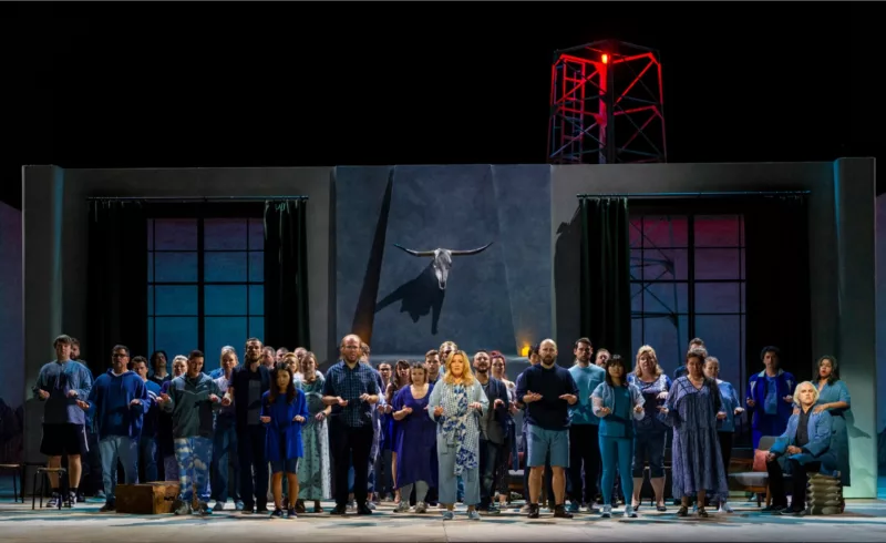 The cast of a new opera having its debut in Philadelphia pose mid-song on stage, in a blue-on-blue wardrobe/set environment, with each person standing face front and stock still,with their arms bent at the elbow and hands out, one palm up and one palm down.