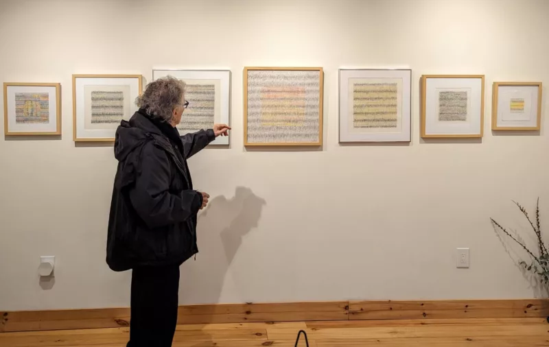 An artist with curly grey hair, wearing a black jacket and slacks stands in a gallery pointing at a piece of framed art on the wall in front of him. He is 83 years old and this is his first solo exhibit and he is talking about his work.