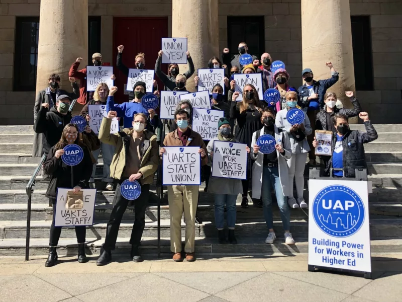 A group of people wearing masks and holding signs supporting a union stand on the steps of a university in Philadelphia.