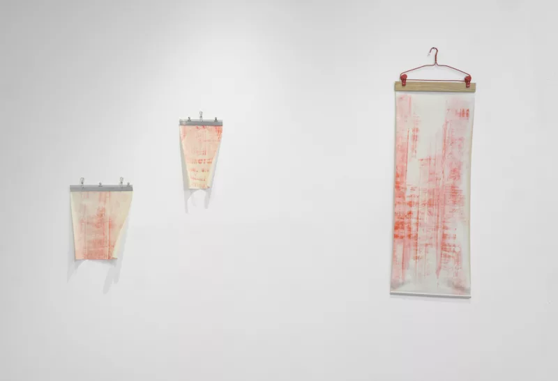 On a white gallery wall, three red and white scroll-like paper works hang together. They are rubbings made of the floorboards in a home.