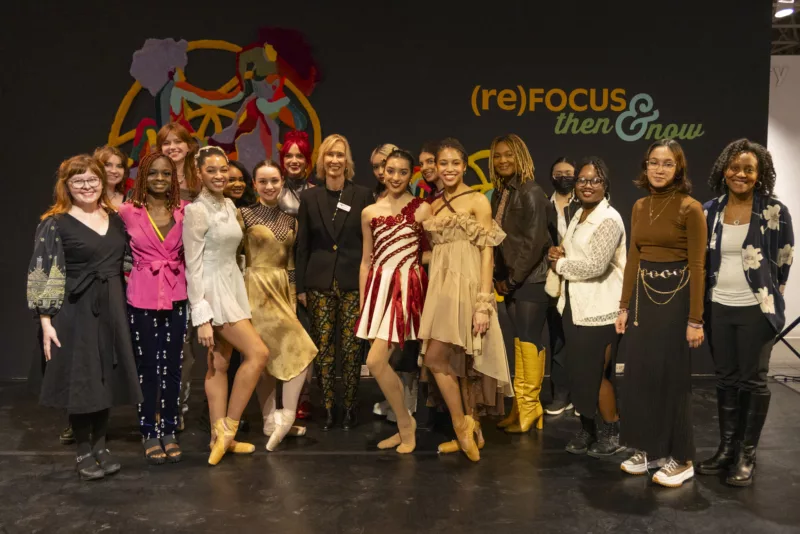 A group of ballet dancers and students and teachers poses in front of a black wall with words on it announcing an art exhibit. The students are juniors at a Philadelphia art school who designed and created the dancers’ costumes. The group is all smiles and proud of their performance and the costumes.