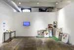 A gallery space with partial interior walls covering a window and duct work in the rear shows a cluster of non-functioning refrigerator doors covered with images perched in a corner on a piece of green astroturf, with a video screen and table to the left.
