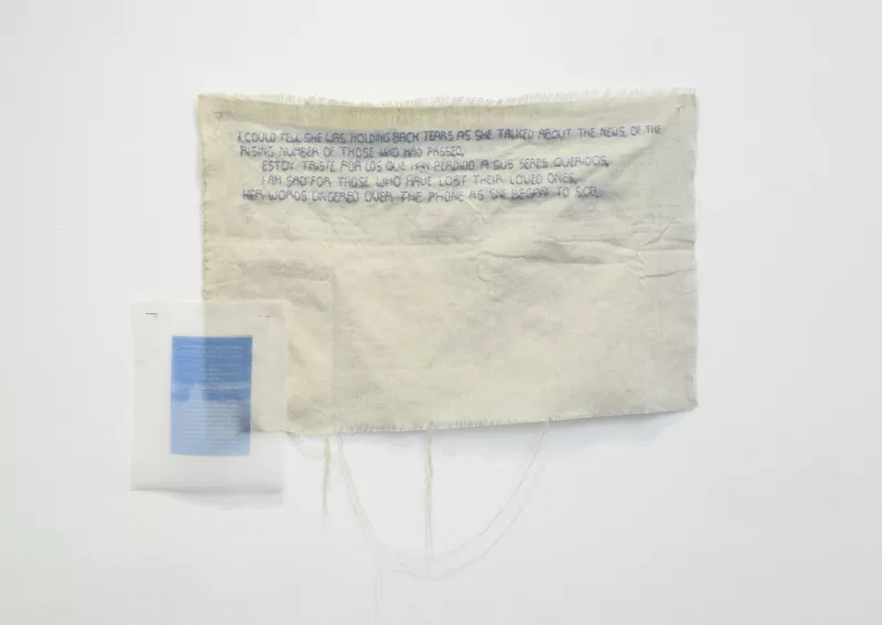 A piece of canvas that is fraying at the bottom, several strings hanging down, has a poem embroidered on it in blue. To the left at the bottom is the poem typed on a blue piece of paper.