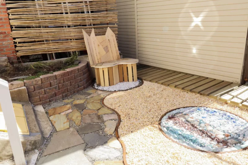 We see the corner of a courtyard, part in shadow, part in sunlight. At the back there’s a hand-made wooden seat. The ground is covered with flagstones and gravel. 