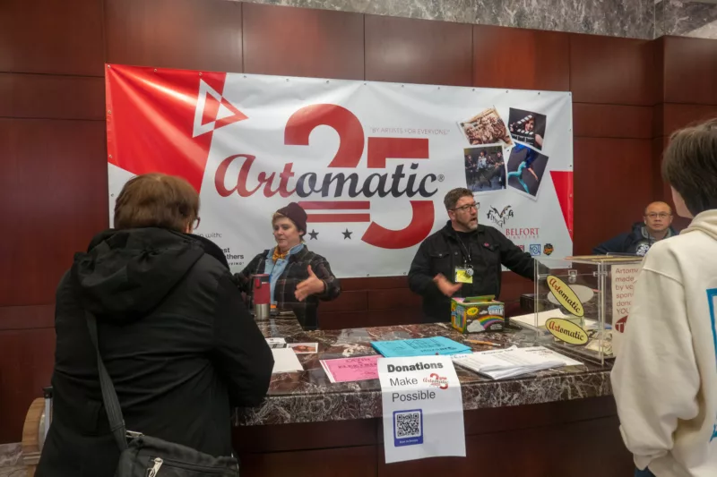 A reception desk in a lobby of Automatic Art Fair shows a big poster on the wall behind two people greeting visitors and a third watching.