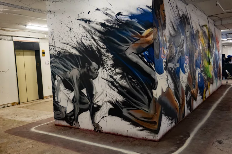 An energetic painted mural showing Black runners in a race wraps a partition wall opposite an elevator in Artomat art fair.