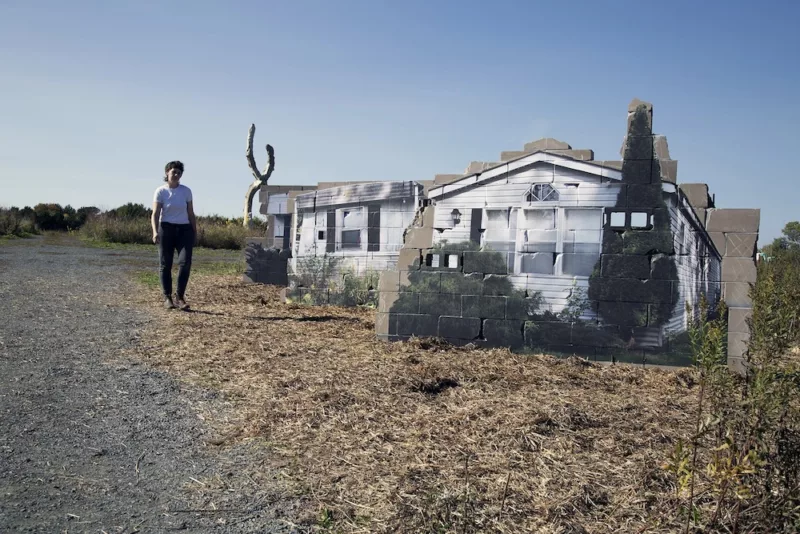 An artist walks in front of several miniature mobile home sculptures situated out in what looks like the middle of a farm field. The artist created the sculptures from used materials.