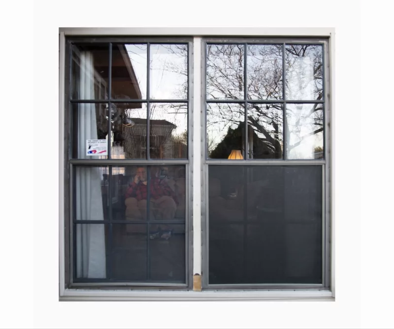 Windows of a mobile home show a senior citizen man sitting on a couch inside. 