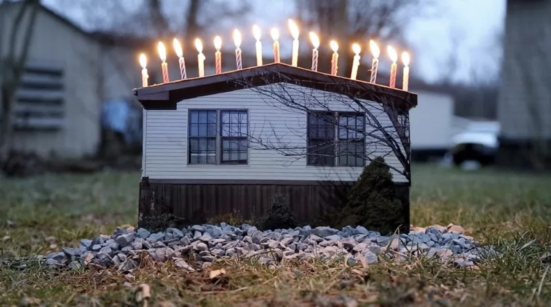 Lit up birthday candles stand like sad soldiers on top of a miniature double-wide mobile home that sits next to a pile of gravel on a grassy plot.