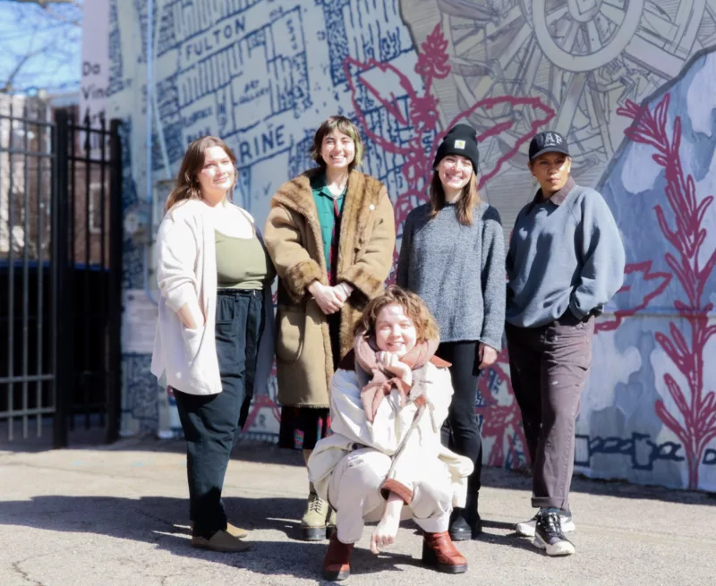 A group of five people, standing in bright sunlight, dressed in winter clothes, stand on the side of DVAA’s building. 