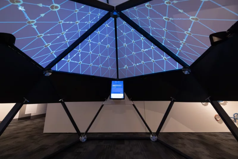 A tent inside a museum has a black armature holding it up and a scrim on the top with a web like print on it. In the tent is a tablet for a participant to ask a question.