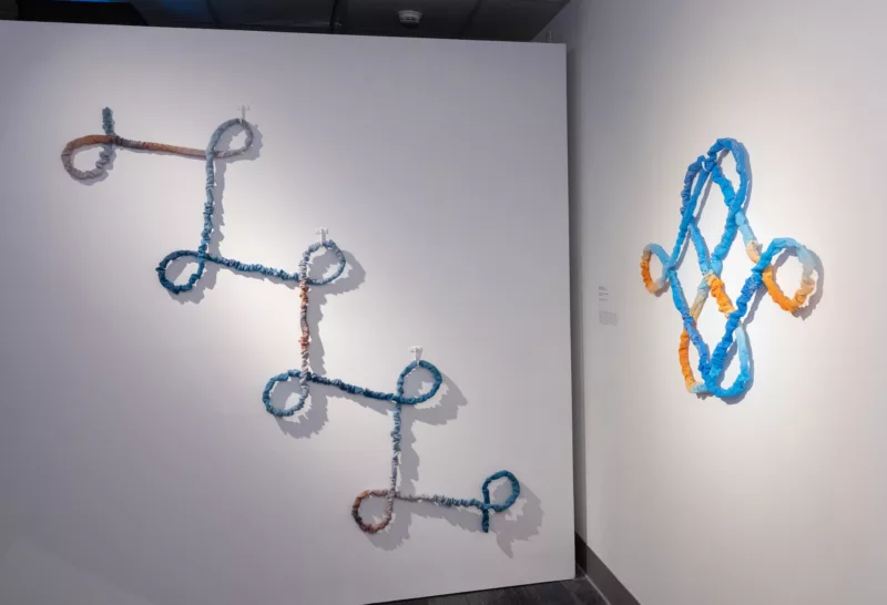 A corner in a museum shows two wall sculptures seemingly made from rope that evoke language and loops. On the left wall a series of cursive “L’s” is linked together, seemingly all made with one piece of rope,