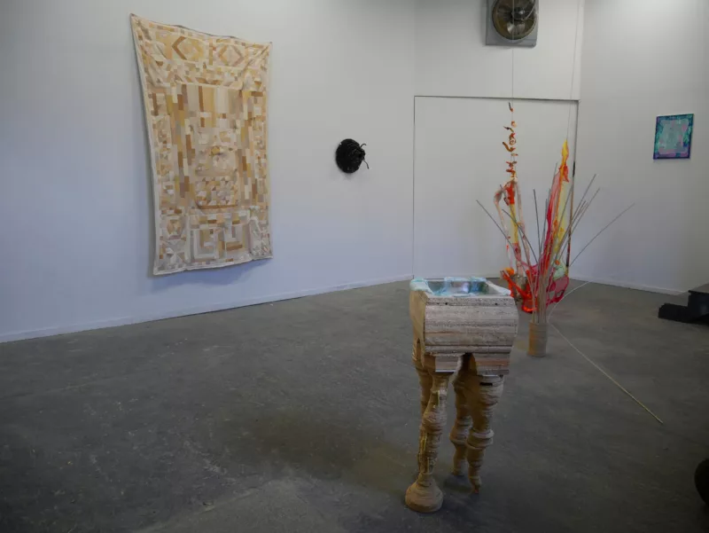 In a gallery with a concrete floor is a group show of works, with several sculptures and some small and large works on the wall. In the foreground is what looks like a basin with four mismatched legs.