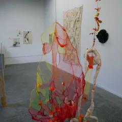 In a gallery with a concrete floor, two colorful and ephemeral sculptures hang close to each other, by strings and chains attached to the ceiling. The right piece seems figural and the left sculpture seems like a net or bag.