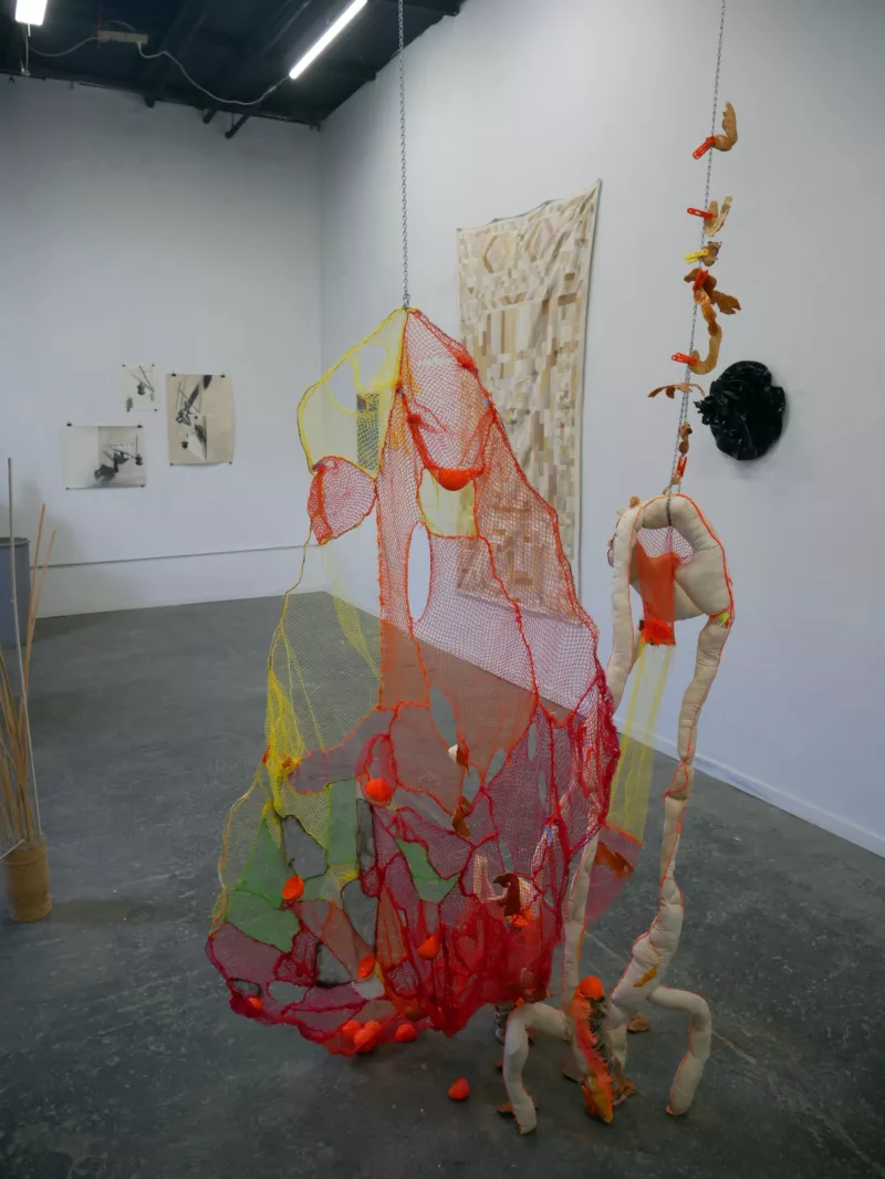 In a gallery with a concrete floor, two colorful and ephemeral sculptures hang close to each other, by strings and chains attached to the ceiling. The right piece seems figural and the left sculpture seems like a net or bag.