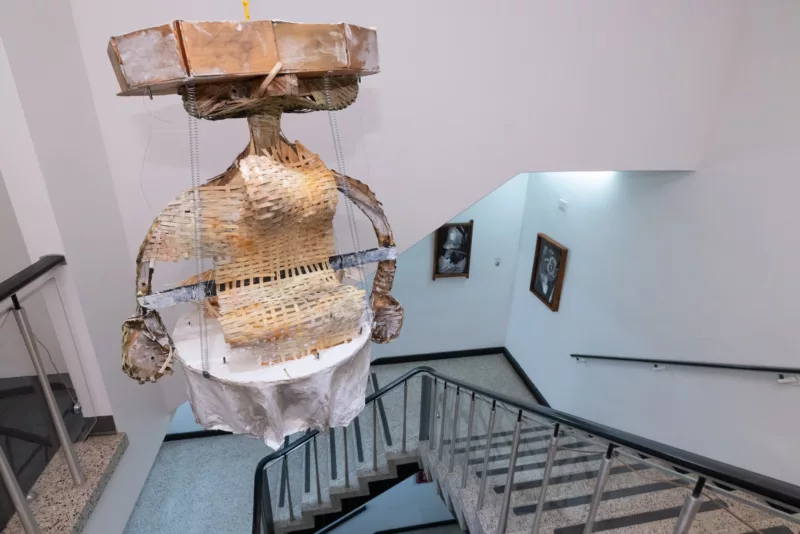 A totemic figure, made of materials like woven paper, cardboard, wood and cloth, hangs in a stairwell, evoking a threat from days of yore when Frankenstein monsters and robots and cyborgs were terrifying images to a general public.