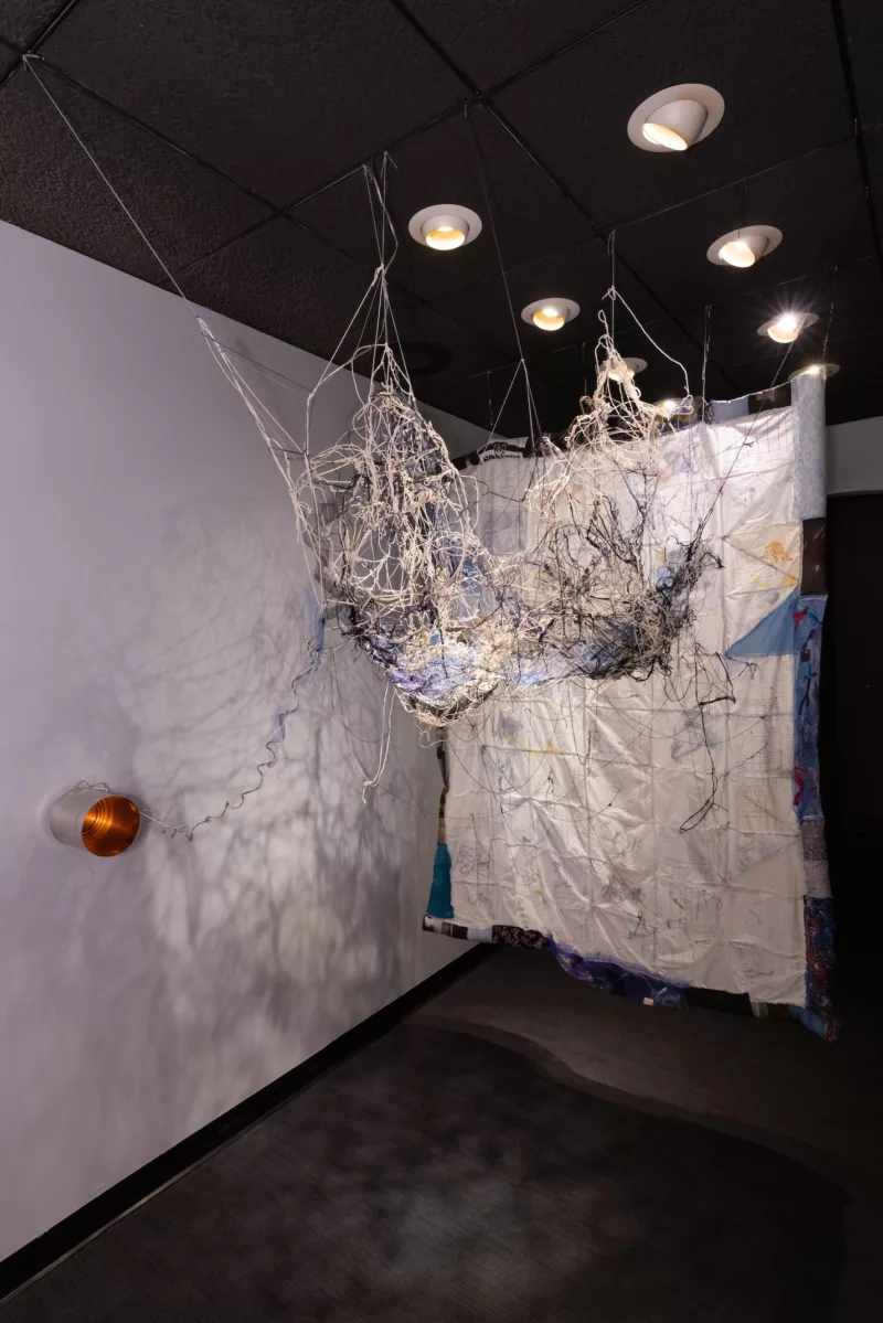 A quilt hangs in a museum space next to rats nest jumble of colored threads of white, beige and black, which are connected to a tin can on the wall its open top towards the viewer. 
