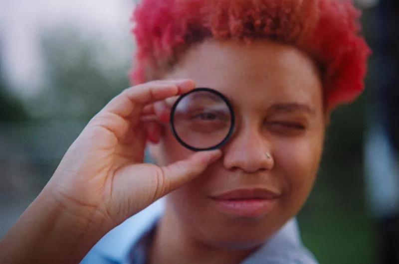 A Black nonbinary person with dyed red hair holds a clear lens up to their right eye and looks through it at you, closing their left eye and smiling. They’re wearing a light blue collared shirt and a stud in their nose. 
