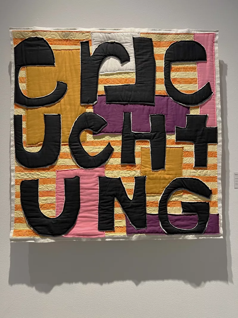 A Colorful quilt with ochre, pink and orange and white striped blocks is the background for bold black cut out quilted letters that are puzzling but spell out the German word, “erleuchtung,” which means, “eureka” or an epiphany.