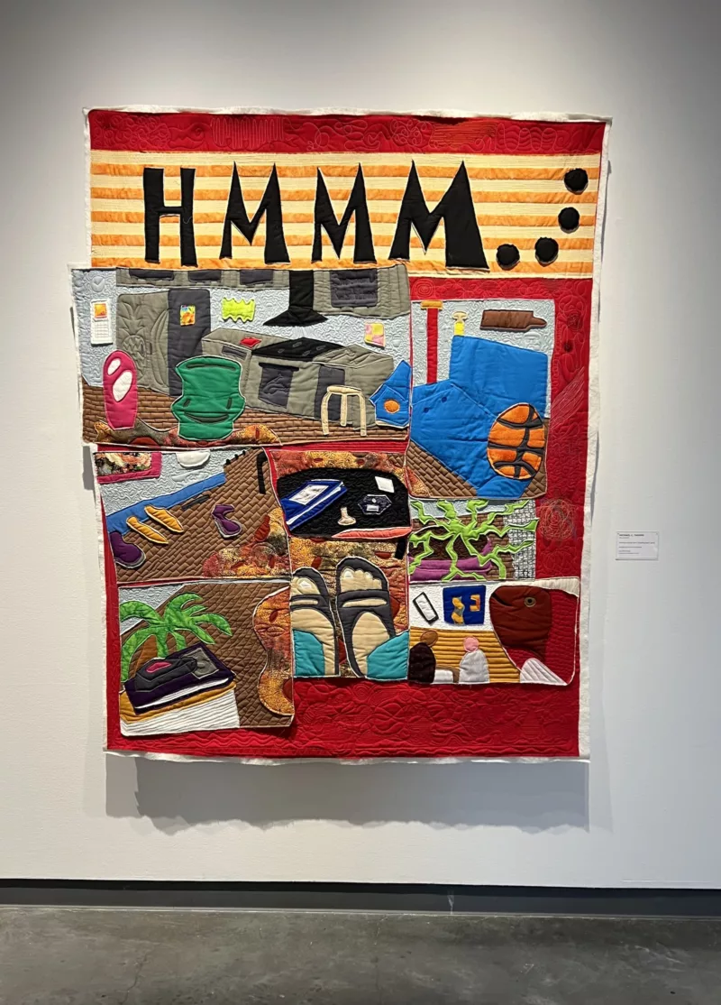 A colorful quilt with the letters “hmmm…” at the top, shows seven separate panels, each depicting a scene from the artist’s home, from shoes lined up on the floor, to chairs and tables, a plant and art on the walls. 