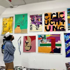 A black male artist, wearing a blue hooded sweatshirt, brown patterned pants and a white baseball hat stands in front of a wall of his quilt works, looking up at them as if studying them before they go off to be in a show.