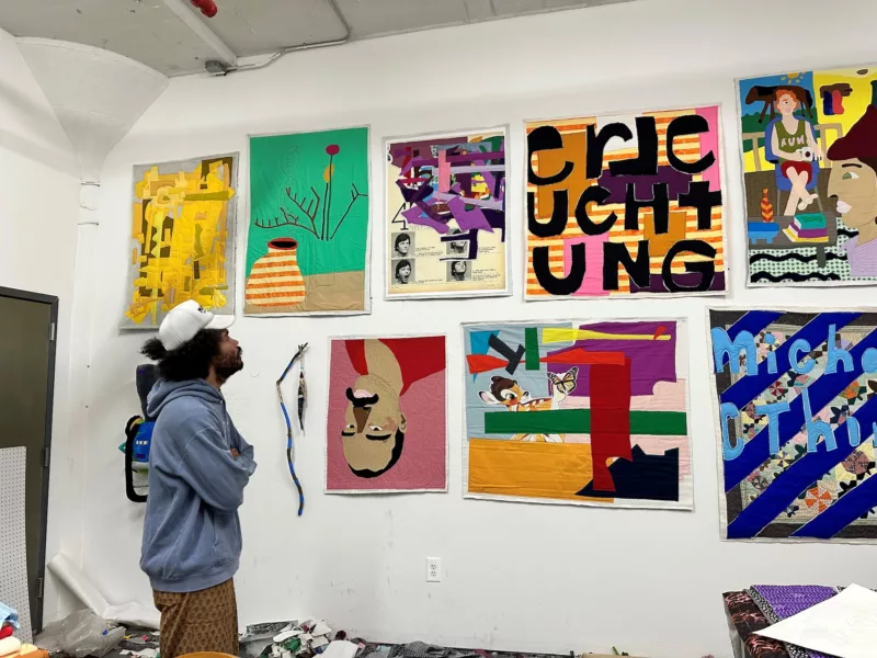 A black male artist, wearing a blue hooded sweatshirt, brown patterned pants and a white baseball hat stands in front of a wall of his quilt works, looking up at them as if studying them before they go off to be in a show. 