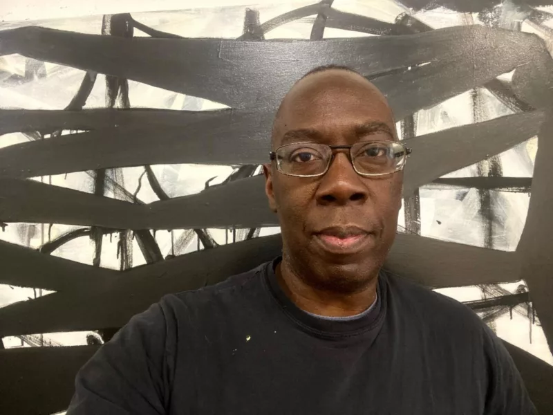 A serious-looking Black man wearing glasses and a black t-shirt stands in front of a black and white painting. 