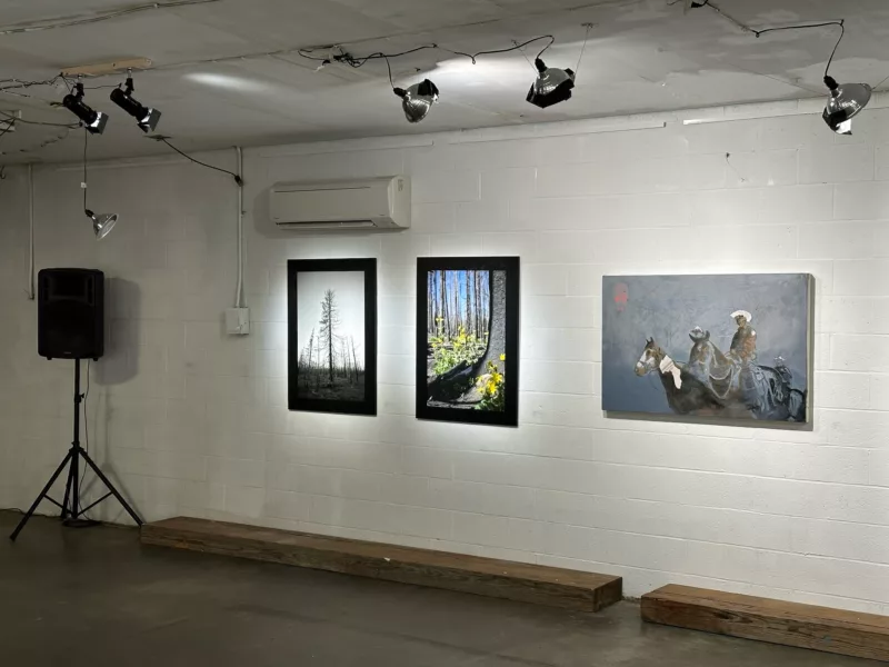 A gallery space shows a white wall with two large color photographs of nature at the left and a painting, very atmospheric, of three cowboys, one a police officer, on a hunt for illegal immigrants at the US border. 