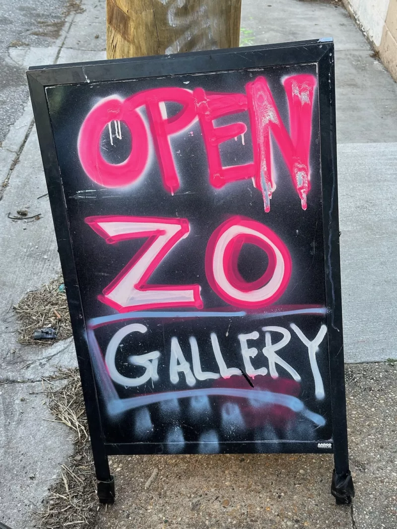 A sign on a city street in Baltimore says “Open, Zo Gallery” with dripping hot pink letters that are graffiti influenced. The gallery is in the western side of Baltimore, Hampden section.