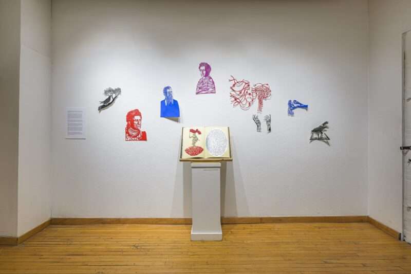 An installation of paper cutouts of a man, a woman, hands, birds, floats on a wall above a pedestal holding an open book. 
