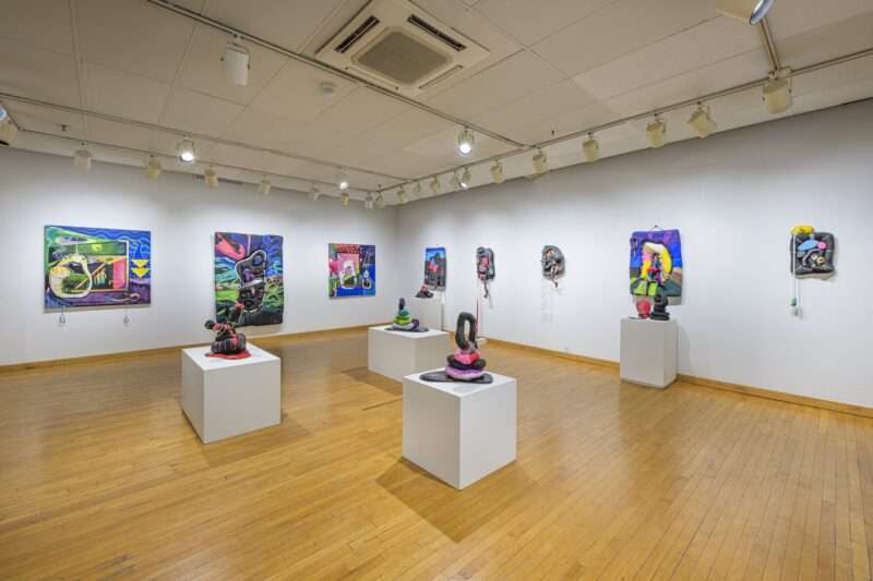 A group of colorful abstract sculptural objects sits on low pedestals in the middle of a gallery, whose walls are covered with likewise bright-colored abstract works in two dimensions. 