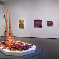 A gallery installation shows four smallish colorful works on a back wall, and in the foreground, on a low white platform, is a large figurative sculpture made from beads and fabric and wood, and metal and found materials, that shows a female figure lying on her stomach with two skinny legs pointing improbably straight up to the ceiling.