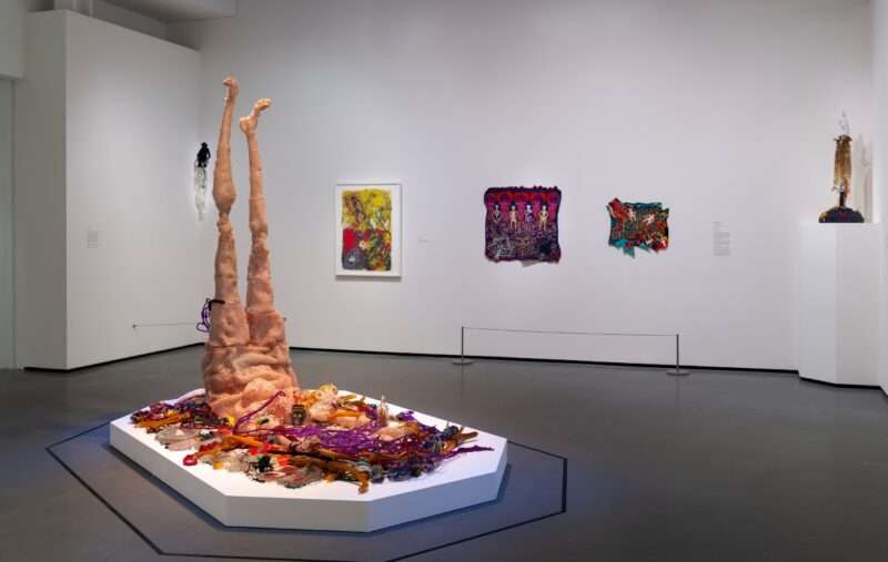 A gallery installation shows four smallish colorful works on a back wall, and in the foreground, on a low white platform, is a large figurative sculpture made from beads and fabric and wood, and metal and found materials, that shows a female figure lying on her stomach with two skinny legs pointing improbably straight up to the ceiling. 