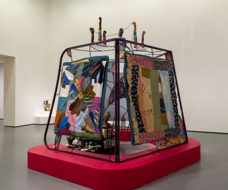 A four-sided metal-framed open structure that evokes a house or dwelling sits atop a low red platform, and on the sides there are quilts fastened to the armature to create a wall or divider. Inside the space is a low altar-like arrangement.