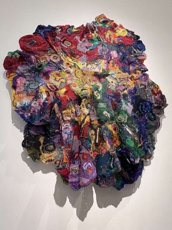 A detailed and tapestry-like wall sculpture with swirls and ribbons of color, beads and knotted fabric; the piece suggests a woman’s hair, seen from the back, perhaps it’s in rows, or it’s an Afro or other stylish arrangement. The piece’s title is “I Call Her Name.”