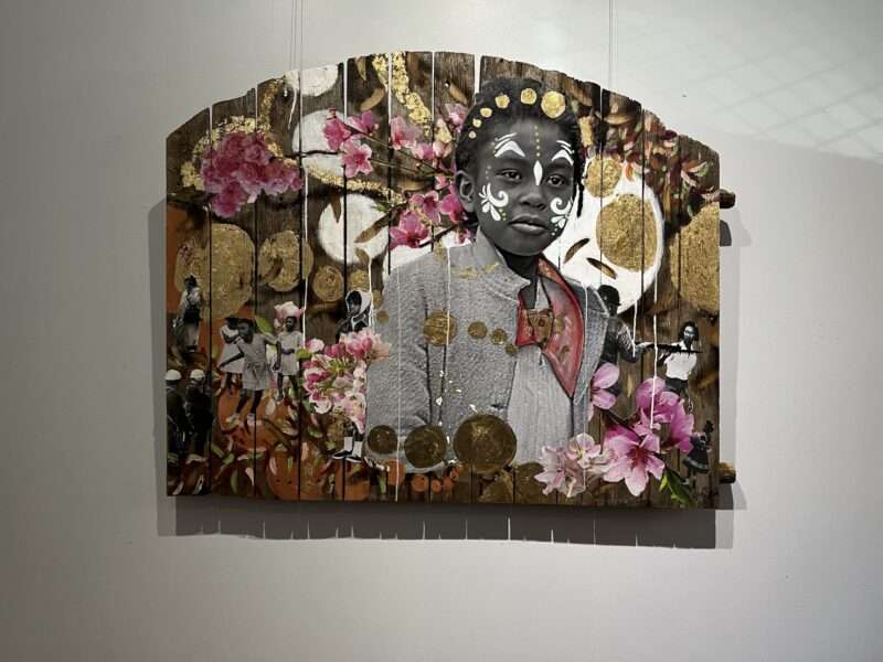 A wall relief made on planks of unpainted picket fencing contains a collaged black-and-white photographic image of a beautiful young Black girl, whose face and hair have been adorned with markings in white and gold circles, that allude to African facial markings and hair adornments. The girl is surrounded by large and small gold circles and flowers.
