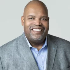 A Black man, who is bald, wears a pale blue shirt and a gray- and blue-checked suit jacket. His teeth gleam white in his big, open smile.