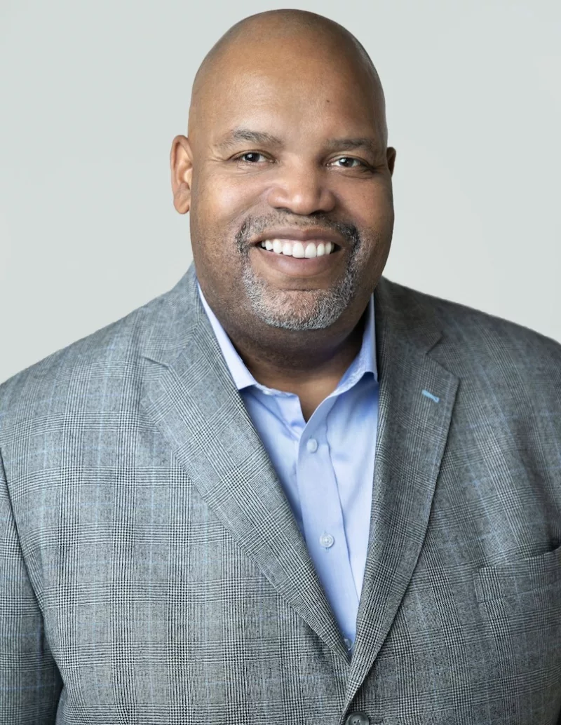 A Black man, who is bald, wears a pale blue shirt and a gray- and blue-checked suit jacket. His teeth gleam white in his big, open smile. 