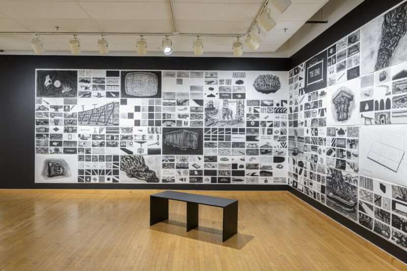 A grid of charcoal drawings spans two walls that meet in the corner of a gallery. A bench sits on the wood floor in front of the large 36 ft. long piece, whose images vary between a magician’s hands, a wall propped up with braces sign that says “The End” and a large nail.