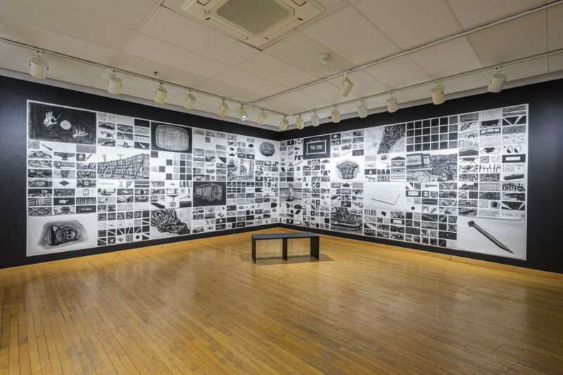 A grid of charcoal drawings spans two walls that meet in the corner of a gallery. A bench sits on the wood floor in front of the large 36 ft. long piece, whose images vary between a magician’s hands, a wall propped up with braces sign that says “The End” and a large nail. 
