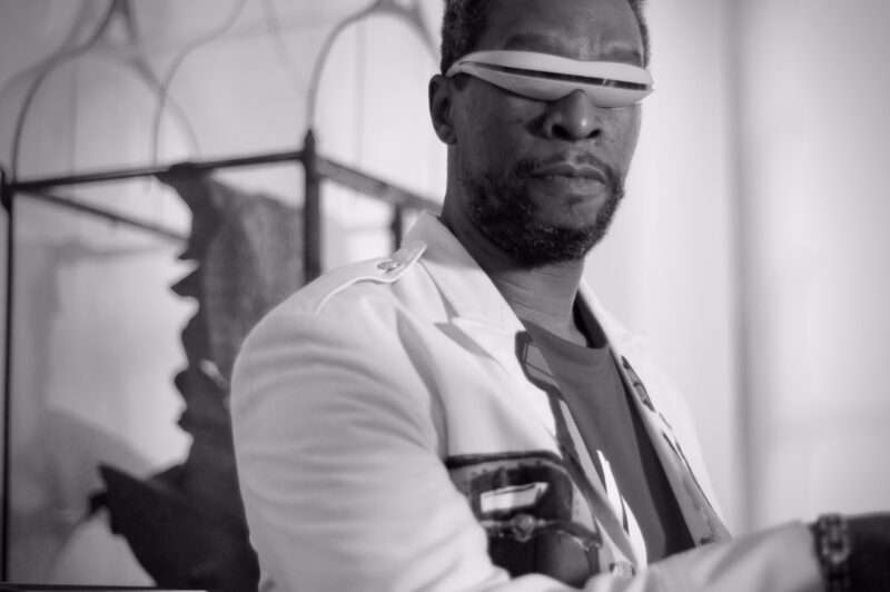 A black and white photo shows a close cropped image of a Black man standing sideways to the camera, with facial hair, wearing a white suit jacket, dark t-shirt and sporting futuristic wraparound sunglasses.